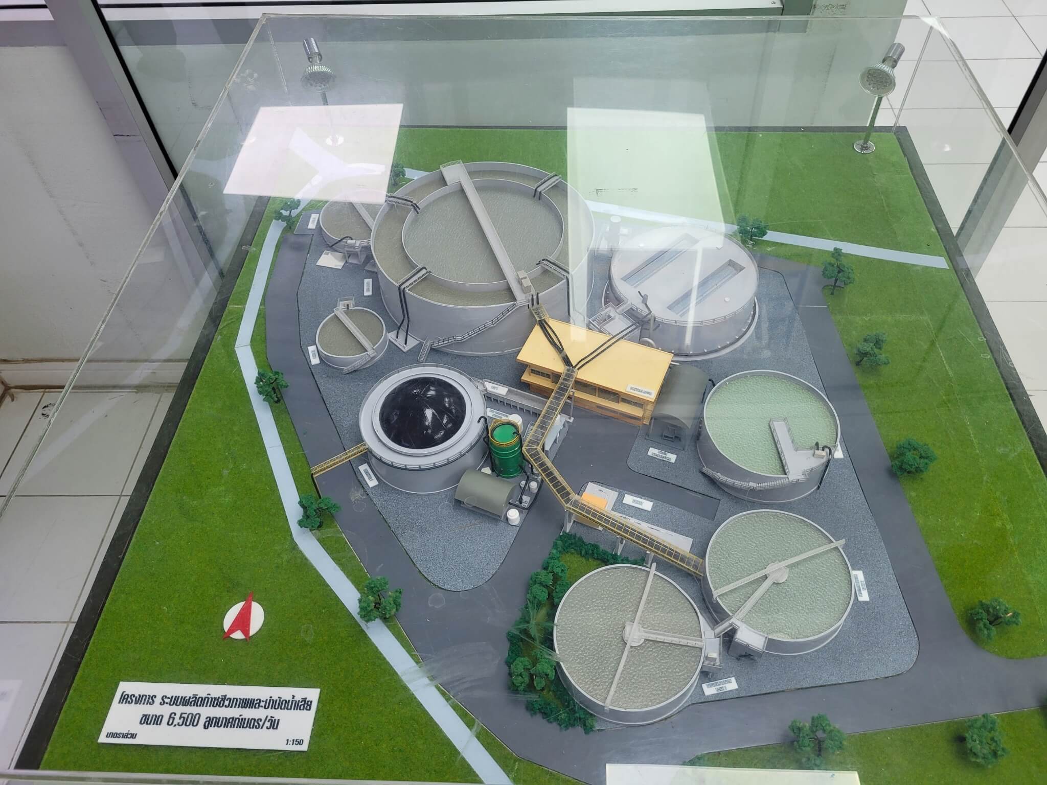 Scale model of a wastewater treatment plant displayed under a glass case, showcasing tanks, pipelines, and facility structures